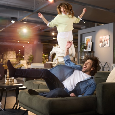 Ein glückliches Pärchen springt vor Freude auf seiner neuen Interliving Couch bei Weser Wohnwelt – die perfekte Kombination aus Komfort und stilvollem Design.